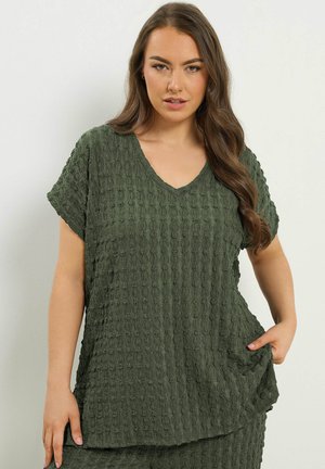 Kvinde med langt bølget hår iført mørkegrøn tekstureret V-hals kortærmet top, holder i sidesømmen, neutral baggrund.