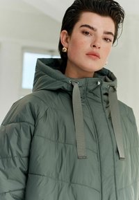 Pūšīgs, kapuces jaka bālā zaļā krāsā, ar horizontālu kvadrātu rakstu, pelēkamiem saites un pogām pie kakla. Gluda, spīdīga tekstūra.