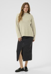 Woman wearing cream ribbed sweater, black midi skirt with front slit, and black loafers, standing against a plain light background.