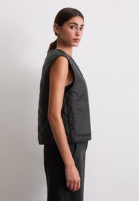 Zwarte gewatteerde gilet met een mouwloos ontwerp, voorzien van een voorzak en een gladde textuur, getoond op een model van opzij.