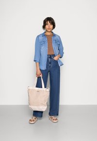Camicia di jeans sopra un top marrone, jeans blu a vita alta e borsa a tracolla beige a righe. Sandali con cinturini completano l'outfit.