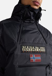 Mustasta nailonista valmistettu vetoketjullinen takki, jossa on korkea kaulus, etupussit ja näyttävä "NAPAPIJRI" -logo, johon on ommeltu Norjan lipun patch.
