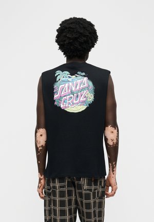 Persona ar tumšiem cirtainiem matiem, valkājot melnu bezroku Santa Cruz krekliņu un rūtainas bikses, stāvot ar muguru un redzamu ādas stāvokli uz rokas.