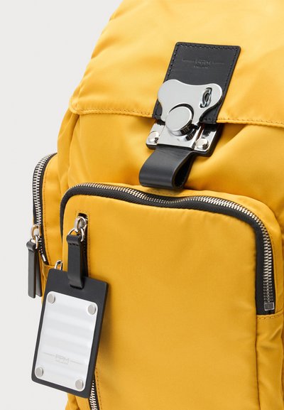 Sac à dos jaune vif avec des fermetures éclair noires, une boucle argentée et une étiquette bagage noire attachée à la poche zippée avant.
