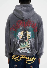 Grå hættetrøje med farverige ulv- og naturgrafikker på ryggen, med "Ed Hardy" tydeligt vist i rød og guld.