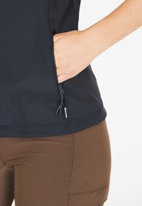 Top nero in tessuto soft-shell con tasca laterale con zip, abbinato a pantaloni marroni attillati. Il tessuto è liscio con una finitura opaca.