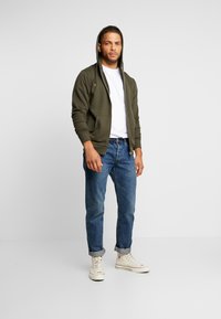 Mann steht da und trägt einen olivgrünen Reißverschluss-Hoodie über einem weißen Shirt, blaue aufgerollte Jeans und weiße High-Top-Sneaker vor einem schlichten Hintergrund.