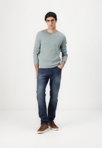 Lichtblauwe gemêleerde trui, donkere spijkerbroek en bruine sneakers. De outfit heeft een ontspannen pasvorm en lange mouwen op de trui.