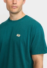 Camiseta de algodón en color verde azulado con mangas cortas, escote redondo y una pequeña cinta de casete bordada en el área del pecho.