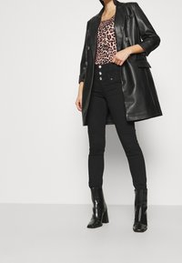 Femme portant un manteau en cuir noir, un haut à imprimé léopard, un pantalon noir taille haute avec des boutons, et des bottines à talons noires.