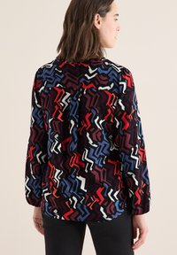 Bluse mit Kragen und einem mehrfarbigen Zickzack-Muster in Rot, Blau, Weiß und Schwarz, mit langen Ärmeln und einer Falte auf der Rückseite.