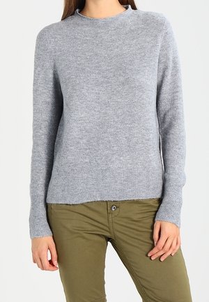 Femme portant un pull en maille gris clair à manches longues et un pantalon vert olive, debout avec les mains détendues le long du corps.
