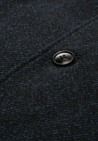 Țesătură din lână închisă, cu un model texturat, având un nasture negru cu patru găuri, poziționat aproape de o cusătură.