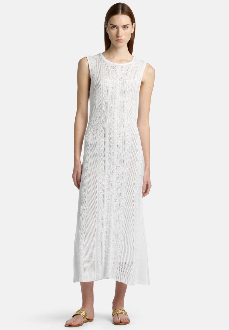 Un vestido maxi blanco sin mangas con un patrón de encaje texturizado; presenta líneas verticales y una silueta fluida. Llevado con sandalias de tono dorado.