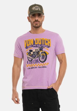 Homme portant un t-shirt violet Von Dutch avec un motif de moto et un short noir, debout avec une main dans la poche et une casquette sur la tête.
