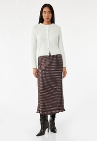 Vit stickad cardigan med knappar, kombinerad med en brun prickig satinmidikjol och svarta knähöga klackstövlar. Slät textur.