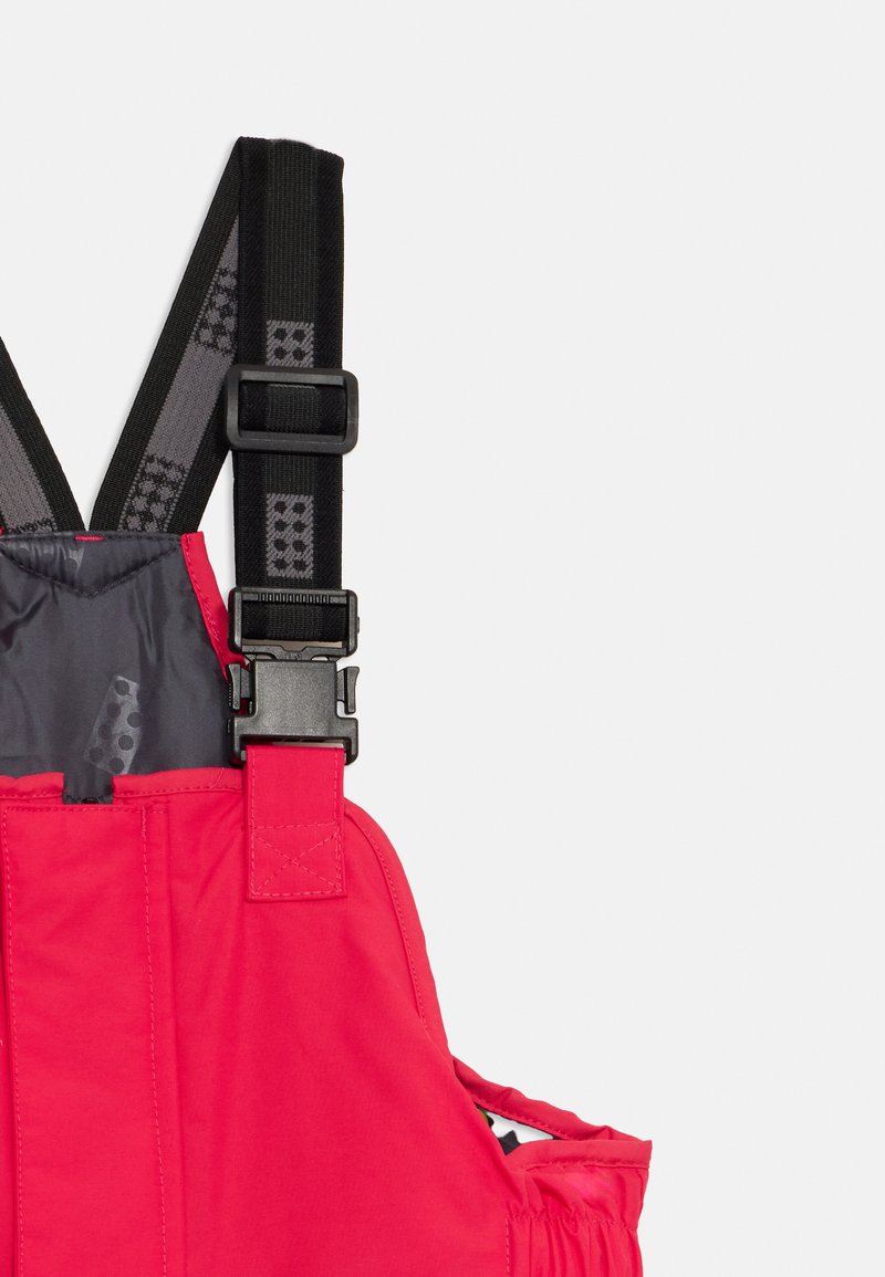 Salopette rossa con bretelle nere regolabili dotate di fibbia in plastica, texture trapuntata e rivestimento interno grigio a contrasto.