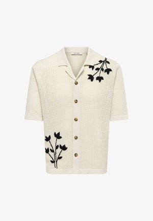 Krém színű rövid ujjú kötött kardigán barna gombokkal, fekete virágos hímzéssel a vállon és az alsó elülső részen, valamint hegyes gallérral.
