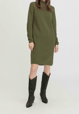 Frau trägt ein langärmeliges, olivgrünes Strickkleid, kombiniert mit schwarzen kniehohen Velourslederstiefeln, steht vor einem schlichten hellen Hintergrund.