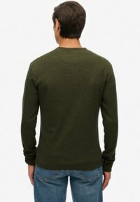 Mand iført olivengrøn langærmet skjorte og blå jeans, stående med ryggen til en ensfarvet hvid baggrund.