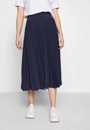 Personne portant une jupe midi plissée bleu marine, un t-shirt blanc et des baskets blanches, debout devant un fond gris uni.