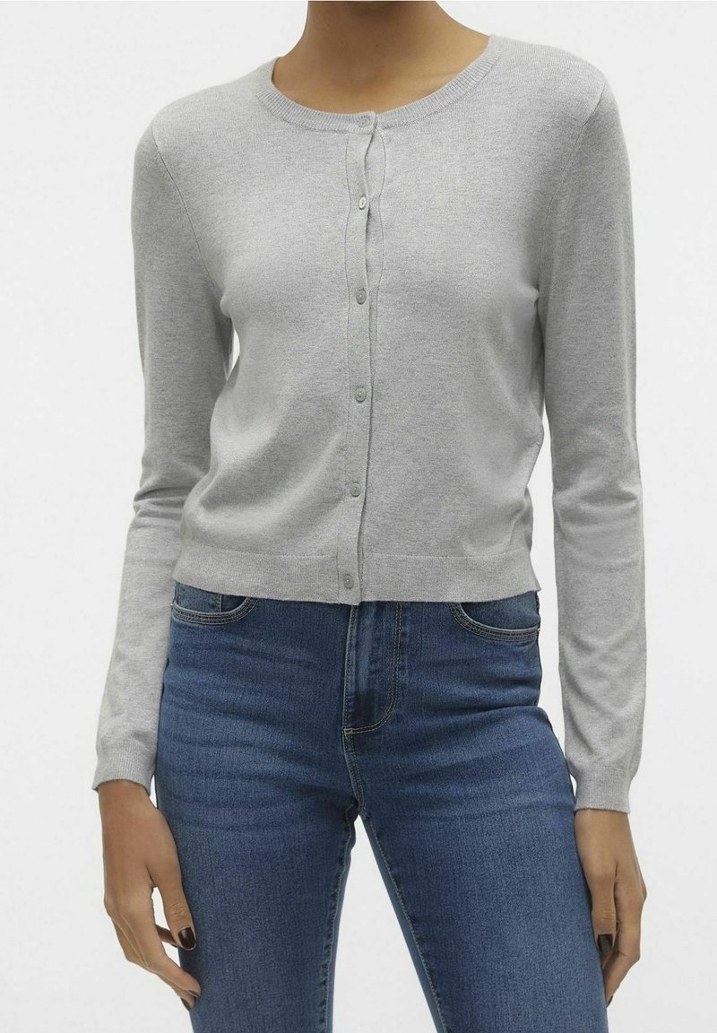 Femme portant un cardigan gris clair boutonné et un jean bleu, mains détendues le long du corps, fond blanc uni.