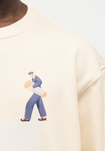 Άνδρας με μπλε στολή που κρατάει ένα μεγάλο δεματισμένο πακέτο με μπαγκέτες, τυπωμένο στο μπροστινό μέρος μιας κρεμ φούτερ.