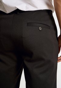 Schwarze Hose mit glatter Textur, tailliertem Schnitt und einer einzelnen Gesäßtasche mit einem schwarzen Knopf. Keine sichtbaren Muster oder Akzente.