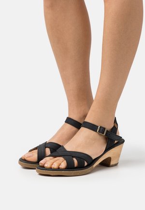 Sandalias de tacón de cuero negro con tiras cruzadas, hebilla ajustable en el tobillo y tacón de cuña de madera. Plantilla texturizada con diseño de punta abierta.