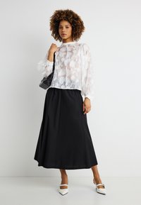 Γυναίκα με σγουρά μαλλιά, φοράει διαφανή λευκή μπλούζα με λουλούδια, μαύρη midi φούστα, λευκά μυτερά παπούτσια και κρατάει μαύρη τσάντα στον ώμο.