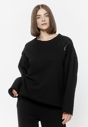 Kobieta o prostych, brązowych włosach do ramion, ubrana w luźną czarną bluzę z detalem nuty muzycznej na lewym ramieniu.