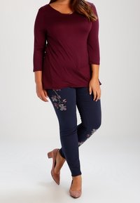 Burgunderfarbener Langarmoberteil kombiniert mit marineblauen Jeans, die mit floralem Stickerei verziert sind. Das Model trägt hellrosa High Heels. Glattes Material mit taillierter Silhouette.