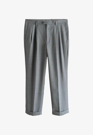 Młody mężczyzna stoi w pomieszczeniu, ubrany w szary dwurzędowy garnitur, białą koszulę, wzorzystą czarną krawat i czarne buty na wzorzystym dywanie.