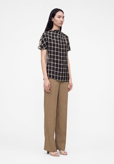 Femme debout portant un haut à manches courtes à carreaux noirs et beiges, un pantalon marron à jambes larges et des talons ouverts beiges sur fond blanc.