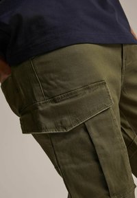 Oliwkowe spodnie cargo z kieszeniami naszytymi, wzmocnionymi szwami i luźnym krojem, zestawione z granatową koszulką.
