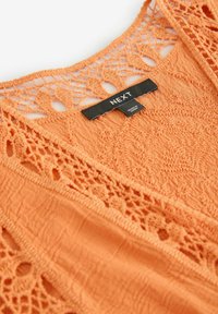 Orange virkade topp med intrikata mönster, en texturerad kropp och en svart etikett med texten "NEXT." Mjuk tyg med öppna vävdetaljer.