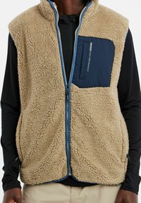 Homme portant un gilet en polaire beige avec une poche poitrine bleue et une fermeture éclair, par-dessus une chemise noire à manches longues et un pantalon beige.
