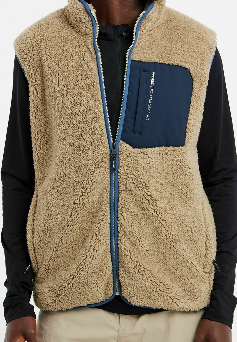 Homme portant un gilet en polaire beige avec une poche poitrine bleue et une fermeture éclair, par-dessus une chemise noire à manches longues et un pantalon beige.