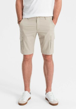 Shorts cargo beiges avec poches latérales, fermeture à bouton et passants de ceinture. Porté avec un t-shirt blanc et des baskets blanches avec des accents bruns.