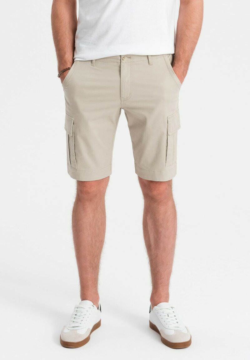 Shorts cargo beiges avec poches latérales, fermeture à bouton et passants de ceinture. Porté avec un t-shirt blanc et des baskets blanches avec des accents bruns.