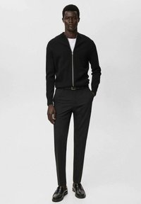 Hombre con un suéter negro con cremallera sobre una camisa blanca, pantalones negros a medida, cinturón negro y zapatos de vestir negros, de pie con una mano en el bolsillo.