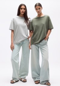 Două modele poartă tricouri oversized: unul în gri deschis și celălalt în verde măsliniu. Ele sunt asociate cu blugi deschiși la culoare cu picioare largi și sandale.