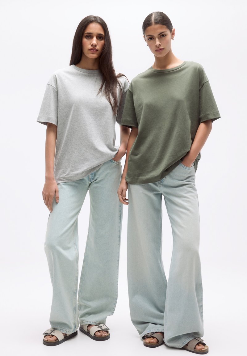 Două modele poartă tricouri oversized: unul în gri deschis și celălalt în verde măsliniu. Ele sunt asociate cu blugi deschiși la culoare cu picioare largi și sandale.