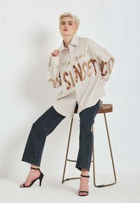 Pruhovaná béžová oversize košeľa s detailom z flitrového nápisu, kombinovaná s tmavomodrými džínsami s rovnými nohavicami a čiernymi sandálmi na podpätku, sediaca na stoličke.