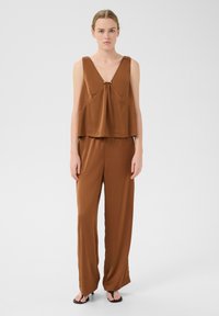 Bronsfärgad ärmlös topp med V-ringning och centrum knutdetalj, kombinerad med vida bronsbyxor i slät tyg. Flip-flop sandals bärs.