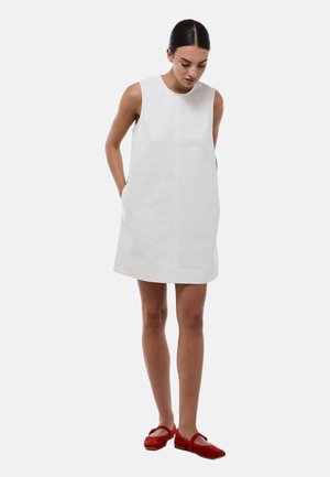 Femme aux cheveux foncés portant une robe droite blanche sans manches et des chaussures plates rouges de style Mary Jane, debout les mains dans les poches, regardant vers le bas.