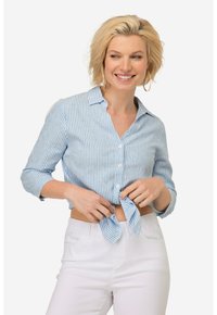 Camicia a righe blu e bianche, corta, con bottoni e colletto, annodata in vita. Indossata con pantaloni bianchi a vita alta.