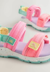 Kleurrijke sandalen met roze stoffen riemen, een paarse voetbed en een witte zool met spikkels, groene accenten en speelse hardware.