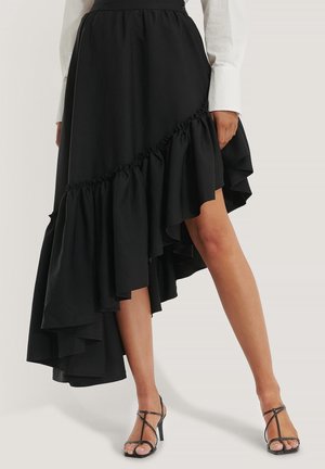 Jupe noire asymétrique à volants portée avec une chemise blanche à manches longues et des sandales noires à talons hauts et à brides, sur jambes nues.