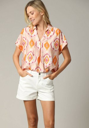 Korte mouw, V-hals blouse met een oranje, roze en witte ikatpatroon. Gecombineerd met witte shorts. Lichte stof, relaxte pasvorm.